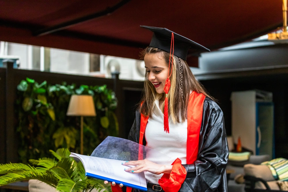 A female graduate in a black and red academic gown looking at her certificate or folder.