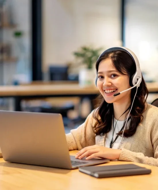 A smiling young woman wearing a headset and working on a laptop in an office setting.
